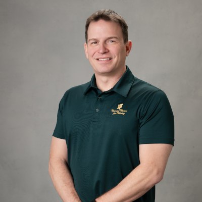 A man with short brown hair smiles while standing in front of a gray background. He is wearing a dark green polo shirt with a small yellow logo on the left side. His arms are relaxed at his sides.