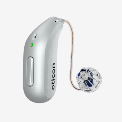 A silver Oticon hearing aid with a slim, curved body, a clear tube, and a dome-shaped earpiece. A small green LED indicator light and button are visible on the side.