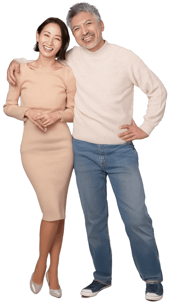 A smiling couple stands together. The woman wears a light beige dress and heels, and the man wears a light sweater, blue jeans, and sneakers. He has his arm around her shoulders. Both look happy and relaxed.